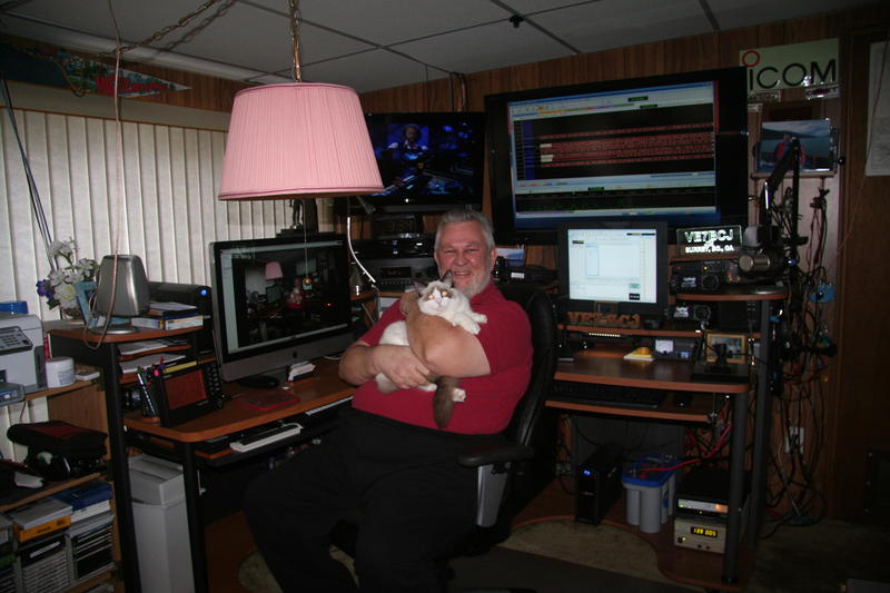 Jim, VE7BCJ, in his radio shack holding his cat, surrounded by Icom and Yaesu radio equipment