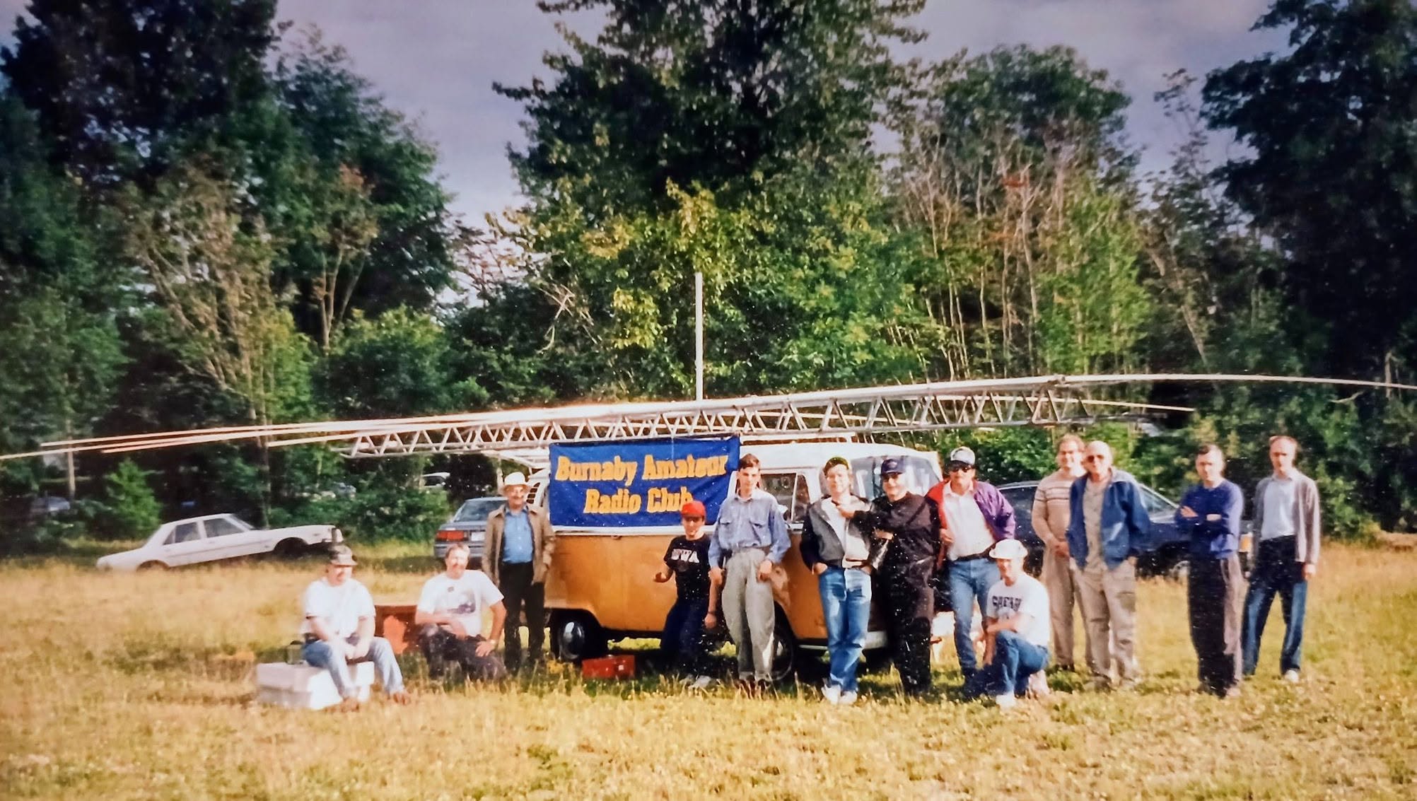 Burnaby Amateur Radio Club members gathered beside Lou Beaubien’s Volkswagen van and a club banner during a Field Day around 1995.